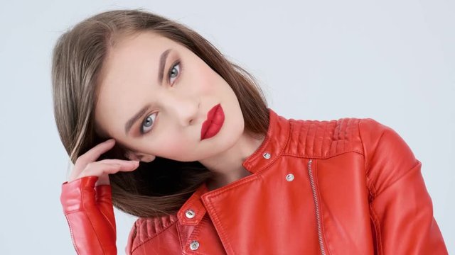 Portrait of a beautiful young woman with red lips. Stylish  girl in a red leather jacket, posing at studio.  Sensual fashion model. Closeup face of a stunning caucasian girl.  4k footage. Slow motion.