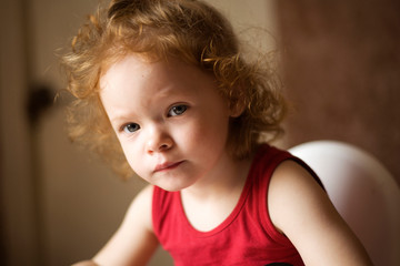 Naklejka premium the little red-haired girl is watching cartoons on the phone , a beautiful baby in a red T-shirt is sitting on the highchair and playing games on the phone portrait