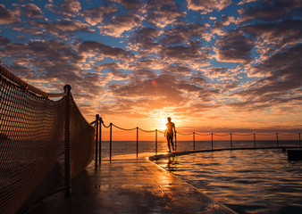 Swimmer walking along the side of Bronte Beach Pool at Sunrise with sunburst. Epic cloudscape.