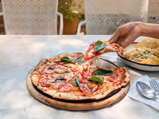 Hand is holding and pulling a slice of pizza margherita out of the tray,happy lunch time with friend in small garden. Foods on white tablecloth.