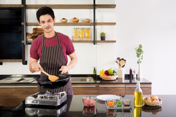 Attractive young man holding pan and wood spatula to make omelette for breakfast at modern kitchen in the morning. New generation lifestyle concept.