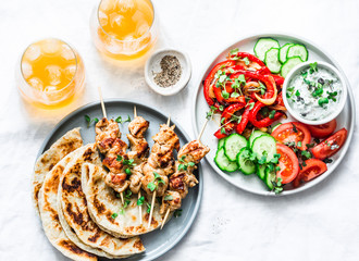 Summer lunch table - turkey skewers, tortillas, tomatoes, cucumber salad, baked sweet pepper, yogurt herb sauce and summer fruit cocktail on a light background, top view