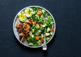 Healthy balanced lunch bowl - turkey skewers, quinoa, avocado, cucumber salad and boiled egg on dark background, top view. Mediterranean style food
