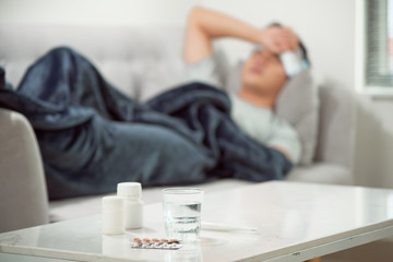 Fototapeta na wymiar sick wasted man lying in sofa suffering cold and winter flu virus having medicine tablets in health care concept looking temperature on thermometer