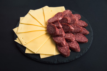 Sliced cheese and salami sausage lie on a round stone black board. Cheese slices on a black background. Sausage sliced on a round board.