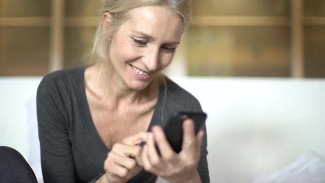 Medium shot of a happy mature woman texting on her smart phone while sitting on a bed