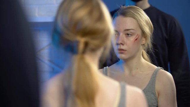 Scared teenager examining face wound mirror, tyrant father standing behind