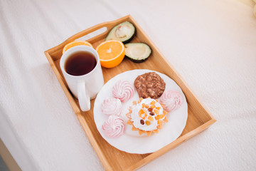 Good morning Breakfast in bed with tea, orange, avocado, cake, marshmallows, chocolate biscuits in wooden tray. lifestyle, food, meal, home comfort and relaxation