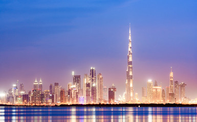 Stunning view of the illuminated Dubai skyline during sunset with the magnificent Burj Khalifa and many other buildings and skyscrapers reflected on a silky smooth water flowing in the foreground.