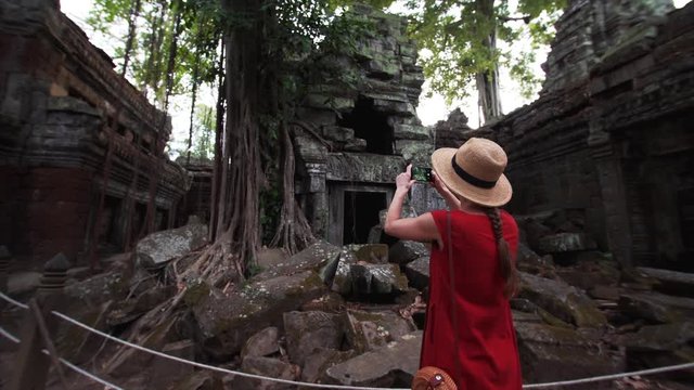 Young woman in straw hat is taking photos of famous Ta Prohm temple ruins and huge banyan roots on them. Angkor Wat complex. Cambodia. UNESCO inscribed Ta Prohm on the World Heritage List in 1992