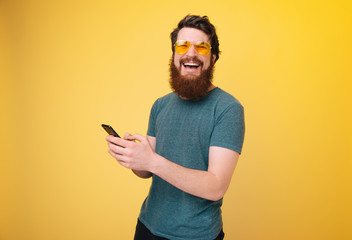 Handsomea bearded guy holding mobile phone and smiling at camera over yllow wall