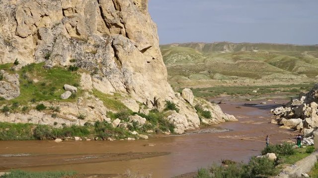 Steady, wide shot of a river below a mountain cliff, man wading in the river with a long pole.