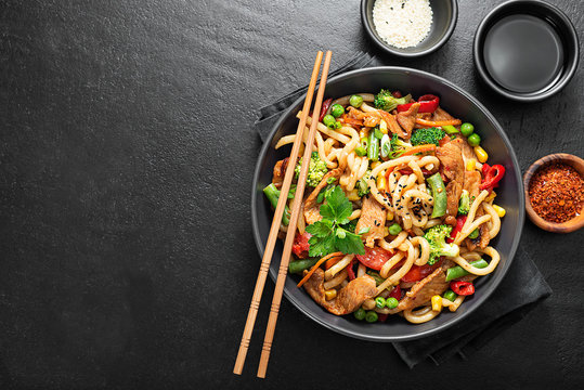 Udon stir fry noodles with pork meat and vegetables in a dark plate on black stone background.