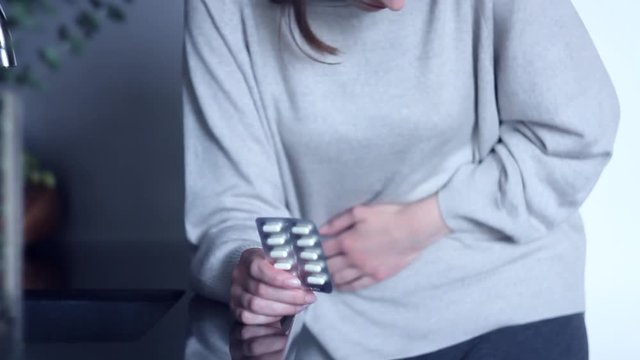 Young woman grabbed her stomach, while standing in kitchen. Girl is holding pills, stroking belly, curving in pain. Sharp attack of pain in abdomen. Disorders and diseases of gastrointestinal tract.