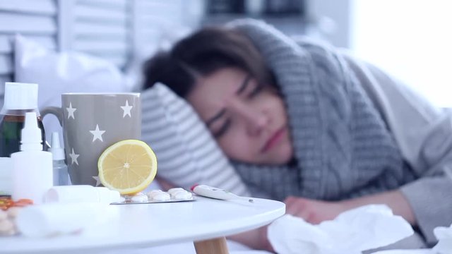 Beautiful sick ill woman female adult with scarf lying on bed with flu at grey bedroom with medicine, drugs and cup of tea with lemon against cold. Coughing and sneezing. Health issue concept.