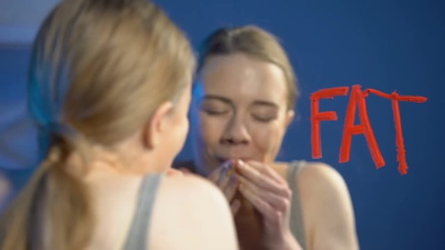 Young woman chewing donut, wiping word fat mirror glass, eating disorder, health