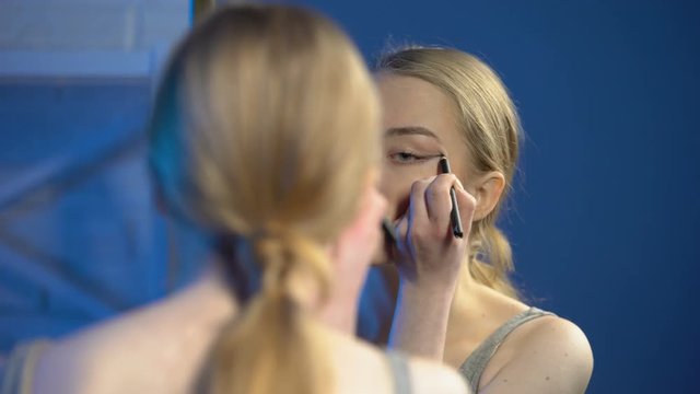 Pretty teenage female applying eyeliner front of mirror, learning to do make-up