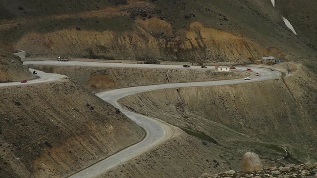 Steady, exterior, aerial, wide shot of vehicles driving on a road winding around mountains.