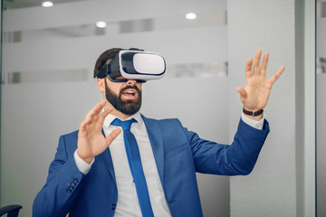 Young Caucasian bearded businessman in blue suit using vr goggles at office.