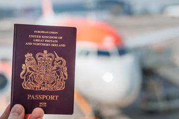 A red British passport held up against a background of a generic plane on a bright sunny day