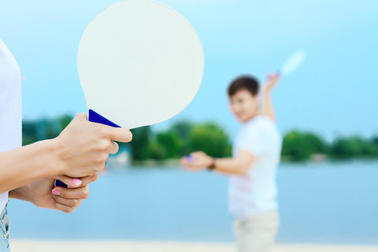 Closeup female hands holding white racket, her smiling boyfriend ready to kick ball on background. Young man and woman in light clothes are playing in matkot on beach. Summer activities concept.