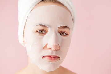 Beautiful young woman is applying a cosmetic tissue mask on a face on a pink background. Healthcare and beauty treatment and technology concept