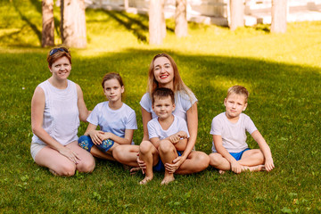 happy women and children sit on the green grass and look at the camera. Big happy family, two mothers and three children in white t-shirts and shorts.