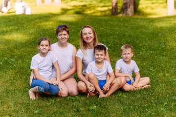 Fototapeta premium Two happy young mothers and three children in white t-shirts sit on a green lawn in the summer. Big Caucasian family on vacation. Women hug children.