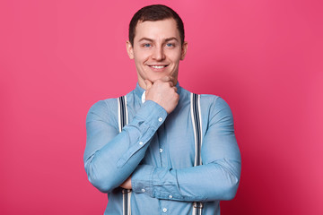 Blue eyed jovial mirthful guy poses in front of camera, touches his chin with one arm, looks delighted, smiles enjoyable. Athletic pleasant man wears blue shirt, white bow tie, striped spenders.
