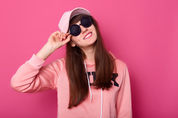 Portrait of happy smiling Caucasian brunette teenager in stylish short pink hoodie, rose cap, black sunglasses, holds hads on hat, ready for going out and spending time with friends. Teenagers concept