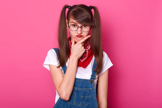 Childish charismatic young lady puts her hand on face, thinking over her plans, makes frowny face. Funny girl with bangs and long pigtails stands straight isolated over bright pink background.