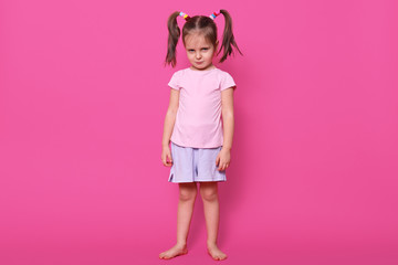 Offended frowned little girl with funny pigtails poses isolated over bright pink background, standing straight and calm, wearing casual light clothes. Childhood concept. Copy space for advertisement.