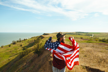 4th of July. Fourth of July. American with the national flag. American Flag. Independence Day. Patriotic holiday. The man is wearing a hat, a backpack, a shirt and jeans. Beautiful sunset light. 