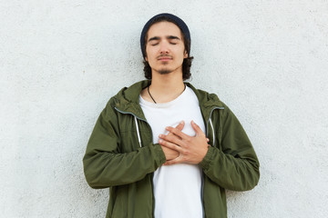 Portrait of sweet handsome youngster leaning on white wall with closed eyes, trying to feel...