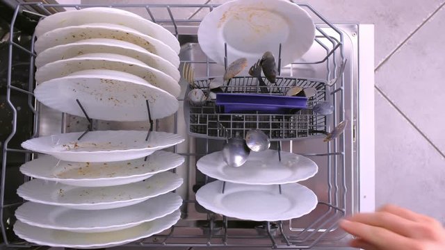 Man's hand puts white dirty plates and spoon in dishwasher and pushes the basket with kitchenware. Top view, close-up hand.