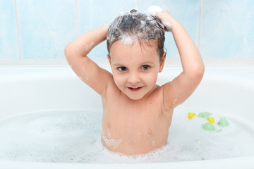 Little cute girl taking bath, washing her hair with shampoo by herself, looks happy playing with foam bubbles, looking directly at camera, infant enjoys being in warm water, charming baby taking bath.