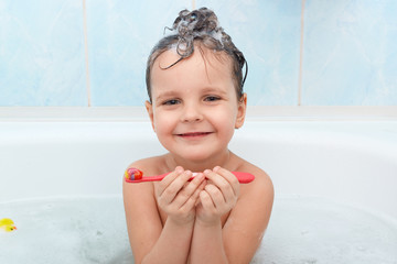 Adorable little baby girl brushing her teeth, taking bath alone, looks smiling at camera, cute little kid glad to wash by herself isolated on blue wall in bathroom. Hygiene, child skin and teeth care.