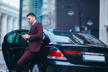 Handsome young businessman leaning on his limo and smiling while looking at his phone.