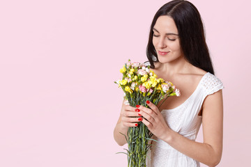 Portrait of tender attractive young woman with long black hair in summer white dress holding bouquet, smelling flowers, isolated over light pink background. People, lifestyle and emotions concept.