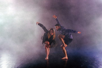 Fototapeta na wymiar Two modern dancers stretching their shoeless feet high in the air surrounded by smoke on stage.