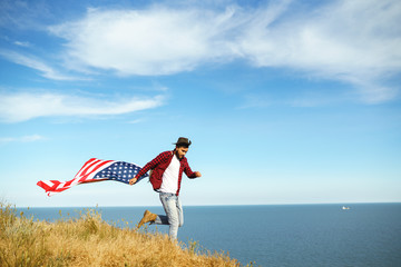 4th of July. Fourth of July. American with the national flag. American Flag. Independence Day. Patriotic holiday. The man is wearing a hat, a backpack, a shirt and jeans. Beautiful sunset light. 