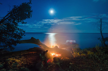 Night fire on the background of the shining moon and glare on the water of the river, lake, sky clouds.