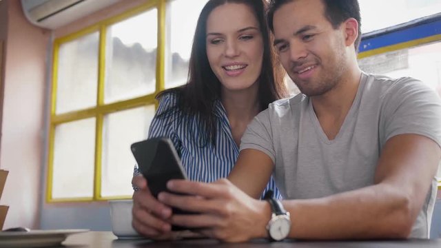 Cute lovely couple enjoying online social life, watching and discussing family photos on smart phone device at cozy loft with window view on the background, scrolling through internet news feed