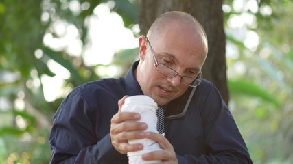 Overworked Young Caucasian Business Man With Two Cups of Coffee Taking Work Phone Call On Lunch Break in Park