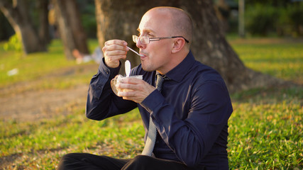 Man Sitting Down, Opening and Eating Yoghurt in Park