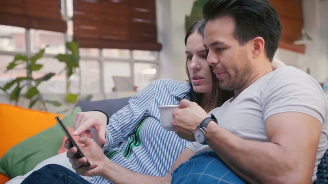 Handsome Hispanic couple lying in cosy living room, using new application on mobile device, doing online shopping on smart phone, chill under warm blue plaid with cup of coffee, wearing watches