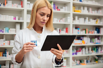 Serious young female pharmacist with blond hair using a tablet to check product description, holding pack of pills in other hand. Shelves with medications in the background