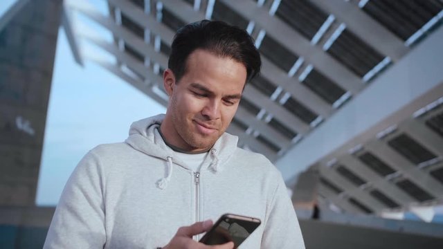 Thoughtful millennial male scrolls news feed in application on the mobile phone device, always staying up to date and online in the internet, looking casual, dressed in grey hoodie, sun battery