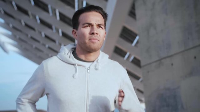 Young attractive Hispanic guy jogging under solar battery panel, doing physical exercises in evening sunlight, running in sportswear, active lifestyle content, concrete building on the background