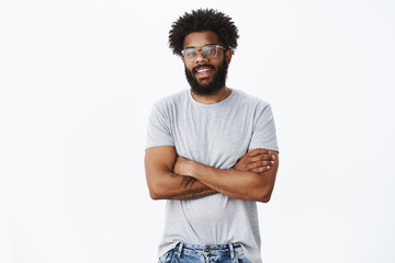Portrait of delighted calm and handsome african american male coworker in glasses with tattoos and pierced nose smiling joyfully and holding hands crossed on chest in confident pose as talking